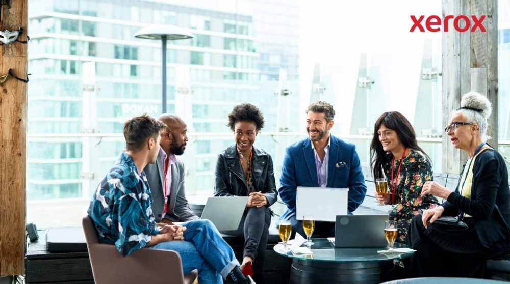 Un grupo de siete personas se sientan juntas, conversan y trabajan con sus computadoras portátiles con una ciudad como telón de fondo. Un grupo de siete personas se sientan juntas, conversan y trabajan con sus computadoras portátiles con una ciudad como telón de fondo.
