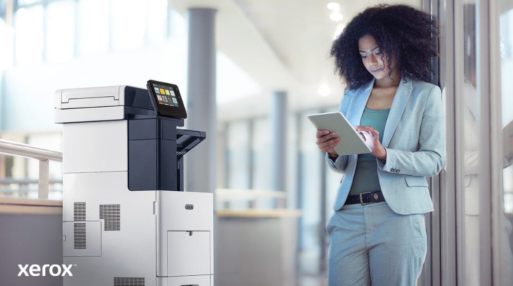 Xerox® ConnectKey®: Conectividad móvil Una persona se encuentra junto a una impresora multifunción Xerox VersaLink B605, mirando e interactuando con una tableta, lo que ilustra cómo ConnectKey permite la interacción móvil con la impresora.