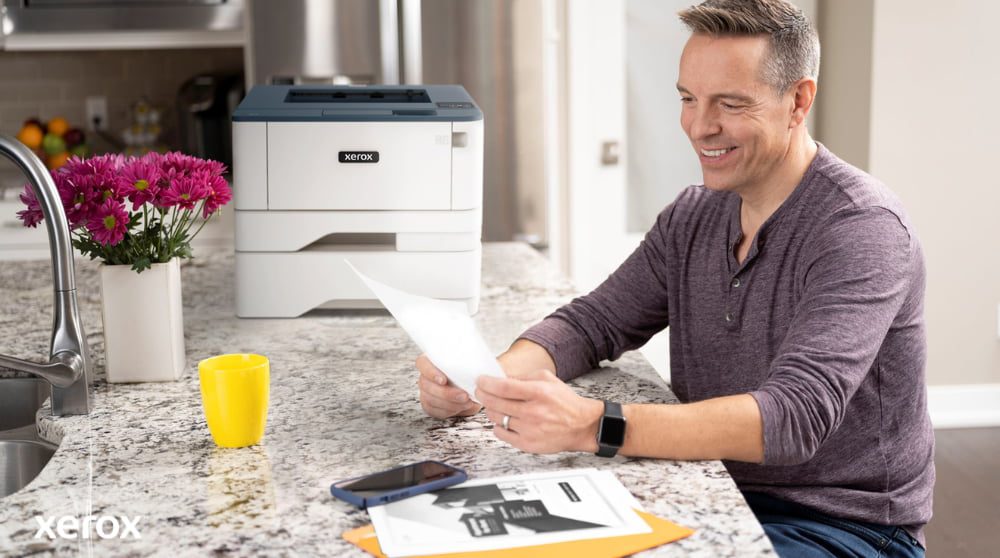 "Experiencia de impresión y escaneo Xerox®: Manejo sencillo de documentos " Una persona sonríe mientras revisa un documento en el mostrador de la cocina, con una impresora Xerox compacta y una taza de color amarillo brillante cerca, lo que sugiere una impresión y un manejo de documentos sin inconvenientes en un hogar o una pequeña oficina.