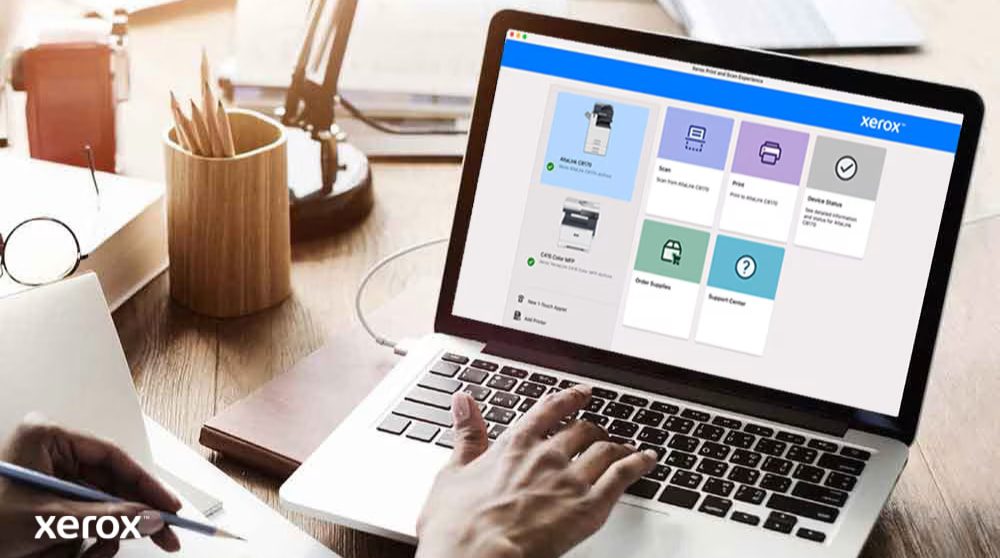 Experiencia de impresión y escaneo de Xerox®: control intuitivo Las manos de una persona están sobre el teclado de una computadora portátil y la pantalla muestra la interfaz de Xerox Print and Scan Experience con opciones como Escanear, Imprimir, Estado del dispositivo, Solicitar suministros y Centro de soporte, lo que indica un control fácil de usar.