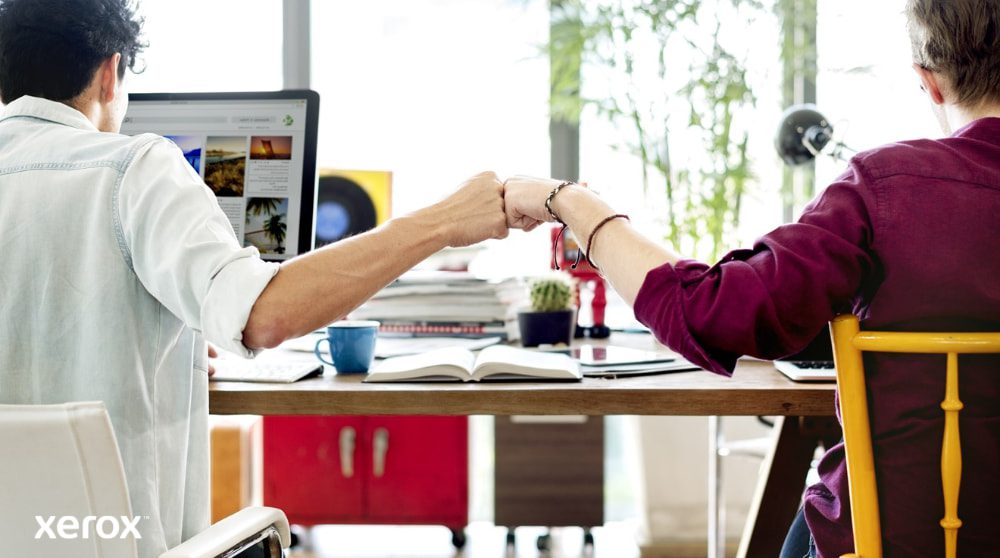 Xerox® Smart Start: Trabajo en equipo y colaboración Dos personas sentadas en un escritorio, de espaldas a la cámara, chocan los puños, símbolo del trabajo en equipo y la colaboración exitosa. Una mira la pantalla de una computadora.