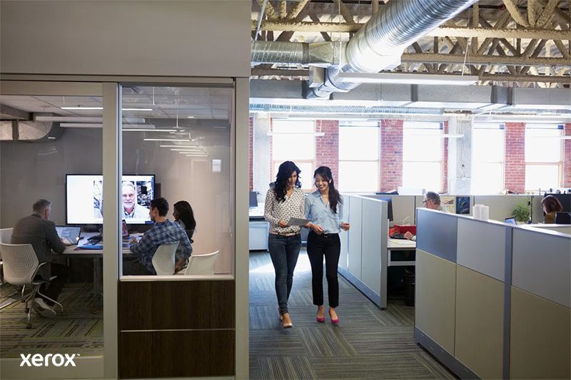 Dos mujeres caminando por un espacio de oficina abierto y moderno, conversando y rodeadas de estaciones de trabajo.
