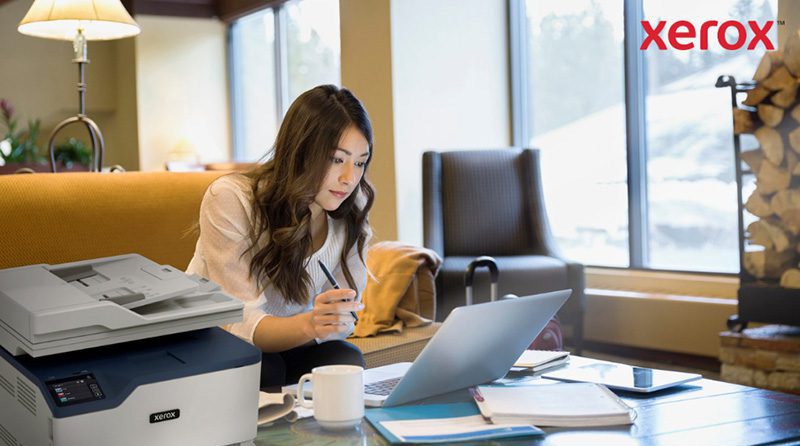 Plataforma Xerox® Workflow Central: Trabaje desde cualquier lugar Una mujer trabaja con una computadora portátil en un cómodo entorno de oficina o sala de estar, con una impresora Xerox compacta visible en primer plano, lo que indica una integración perfecta de la impresión con los flujos de trabajo digitales.