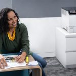 Impresora Xerox® VersaLink® C620 en una oficina Mujer sonriente en una oficina junto a la impresora a color Xerox® VersaLink® C620