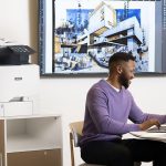 Impresora multifunción en color Xerox® VersaLink® C415 en una oficina Hombre trabajando con un portátil delante de una impresora multifunción en color Xerox® VersaLink® C415